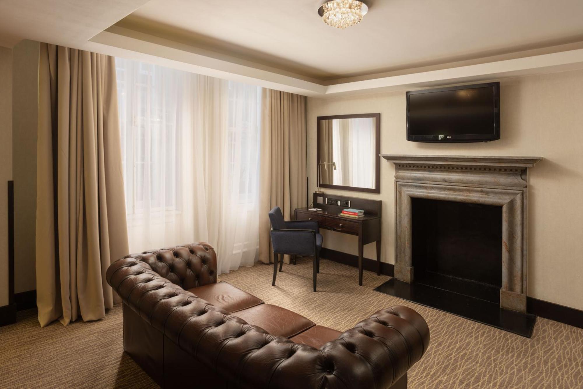 Montcalm Brewery Townhouse Hotel Londra Exterior foto The photo shows a stylish interior of a room, likely a living area or a hotel suite. It features a dark leather chesterfield sofa with tufted upholstery. Opposite the sofa is a small wooden desk with a chair, positioned near a wall-mounted flat-scree