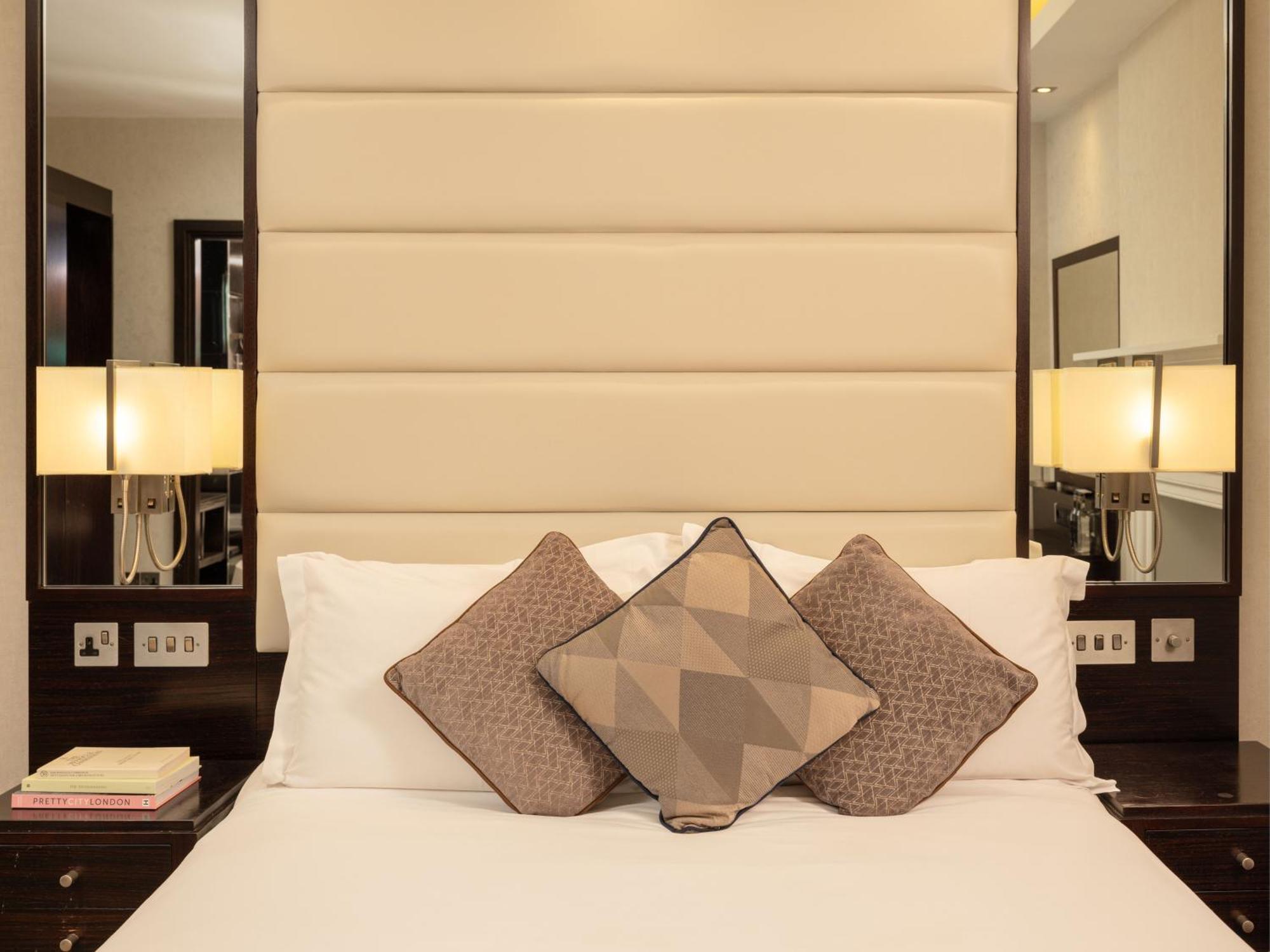 Montcalm Brewery Townhouse Hotel Londra Exterior foto The image shows a cozy bedroom setting. The bed features a headboard upholstered in a light beige fabric. It is adorned with three decorative pillows in varying patterns and colors, placed on white bedding. On either side of the bed, there are modern