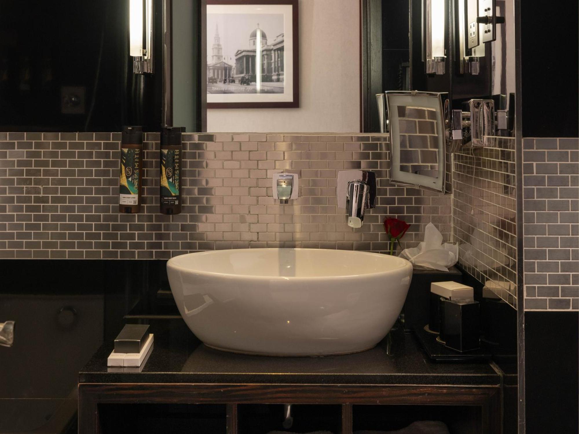 Montcalm Brewery Townhouse Hotel Londra Exterior foto The photo shows a modern bathroom with a stylish design. It features a large, round white sink placed on a dark wooden vanity. The walls are adorned with a mix of gray and silver tile, creating a sleek and contemporary look. Above the sink, there is 