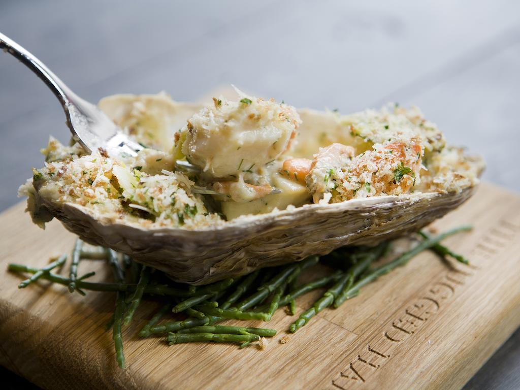 Montcalm Brewery Townhouse Hotel Londra Exterior foto The photo shows a large oyster shell filled with a mixture that appears to include seafood, herbs, and breadcrumbs. The dish is presented on a wooden board with some greenery, possibly a type of seaweed or garnishing. A fork is placed beside the oyst