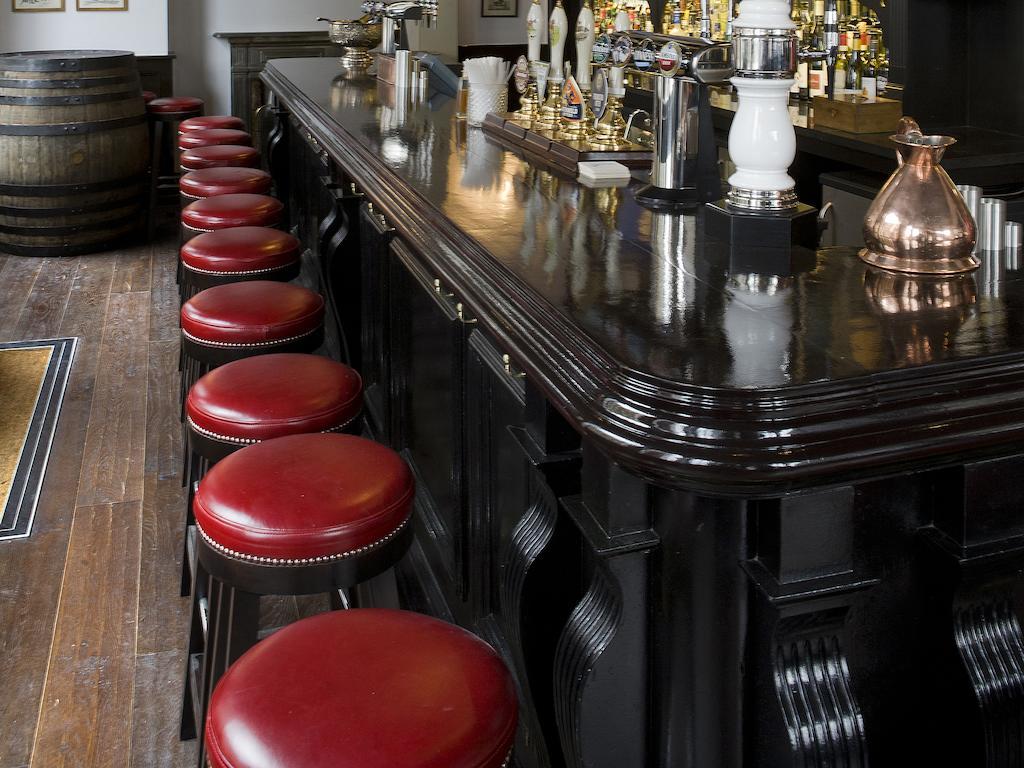 Montcalm Brewery Townhouse Hotel Londra Exterior foto The photo shows a bar setting with a long, polished dark wooden bar counter. Attached to the counter are several bar stools with red leather seats. Behind the bar, there are various bottles, suggesting a well-stocked bar. The flooring appears to be w