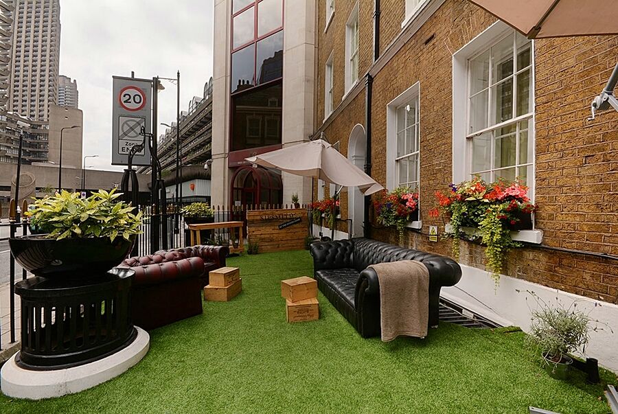 Montcalm Brewery Townhouse Hotel Londra Exterior foto The photo depicts a charming outdoor space, likely a patio or garden area. It features two comfortable-looking leather sofas, one of which is a deep burgundy color, placed on a green artificial lawn. There are wooden crates that could serve as side t